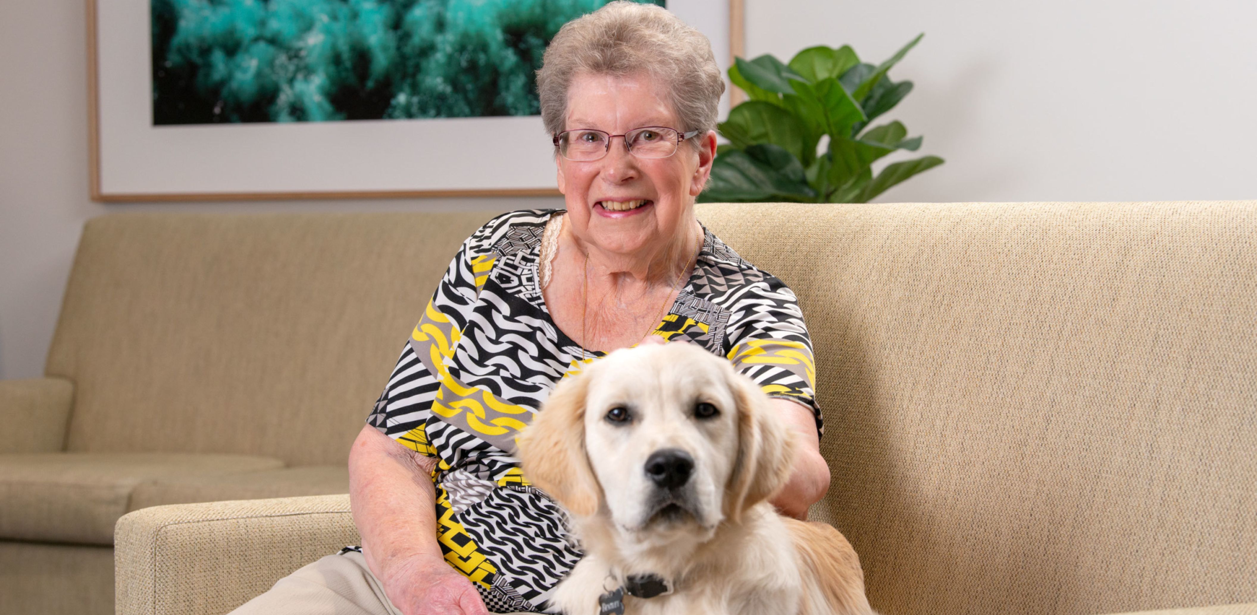 An Eldercare resident smiles at the camera as she sits on an armchair with a golden retriever puppy sitting beside her.