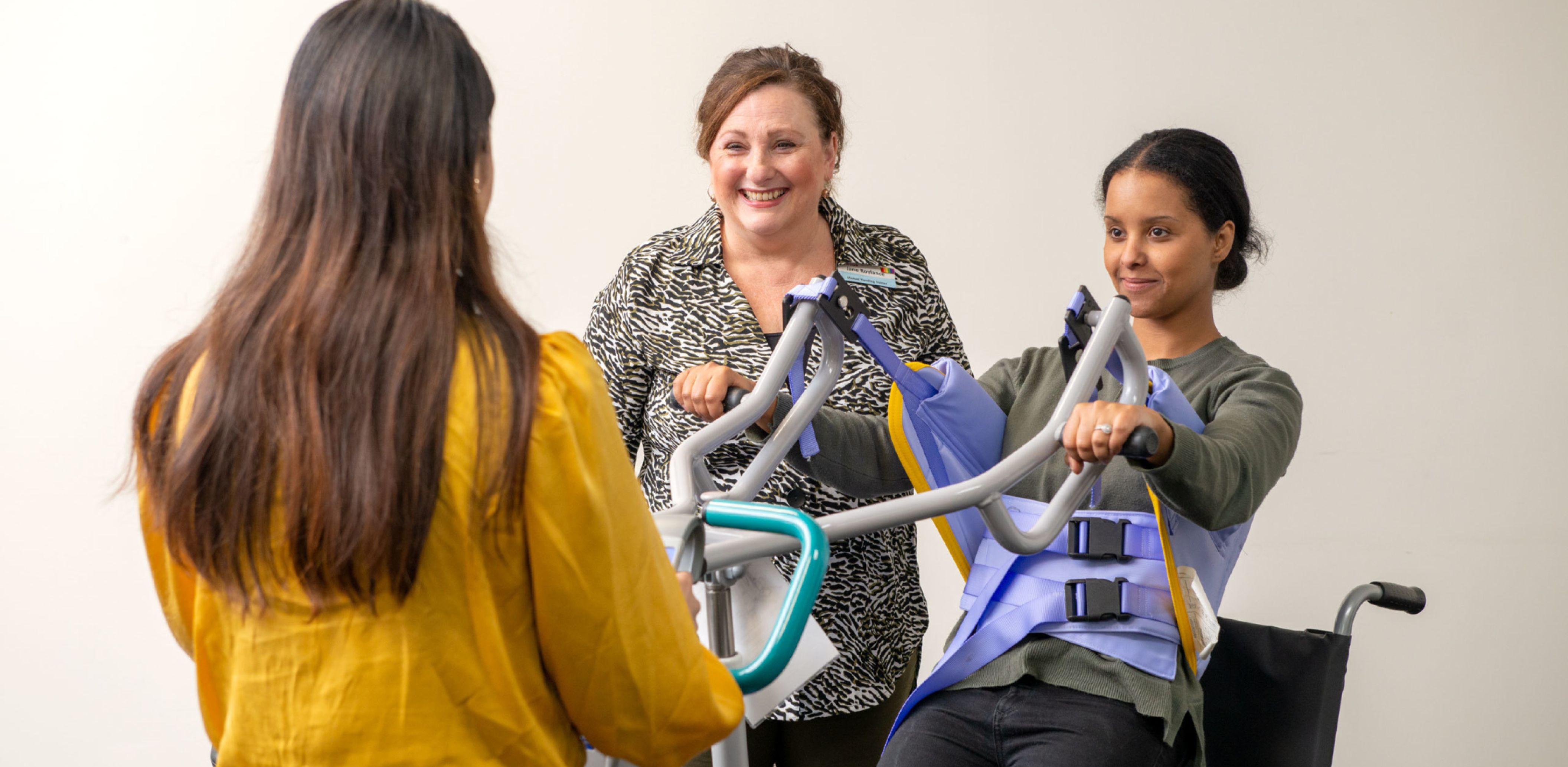 Two Eldercare staff members operate a lifter while the trainer looks on and smiles off-camera.