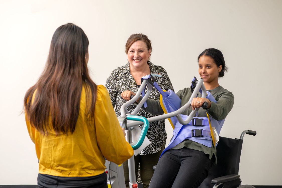 Two Eldercare staff members operate a lifter while the trainer looks on and smiles off-camera.