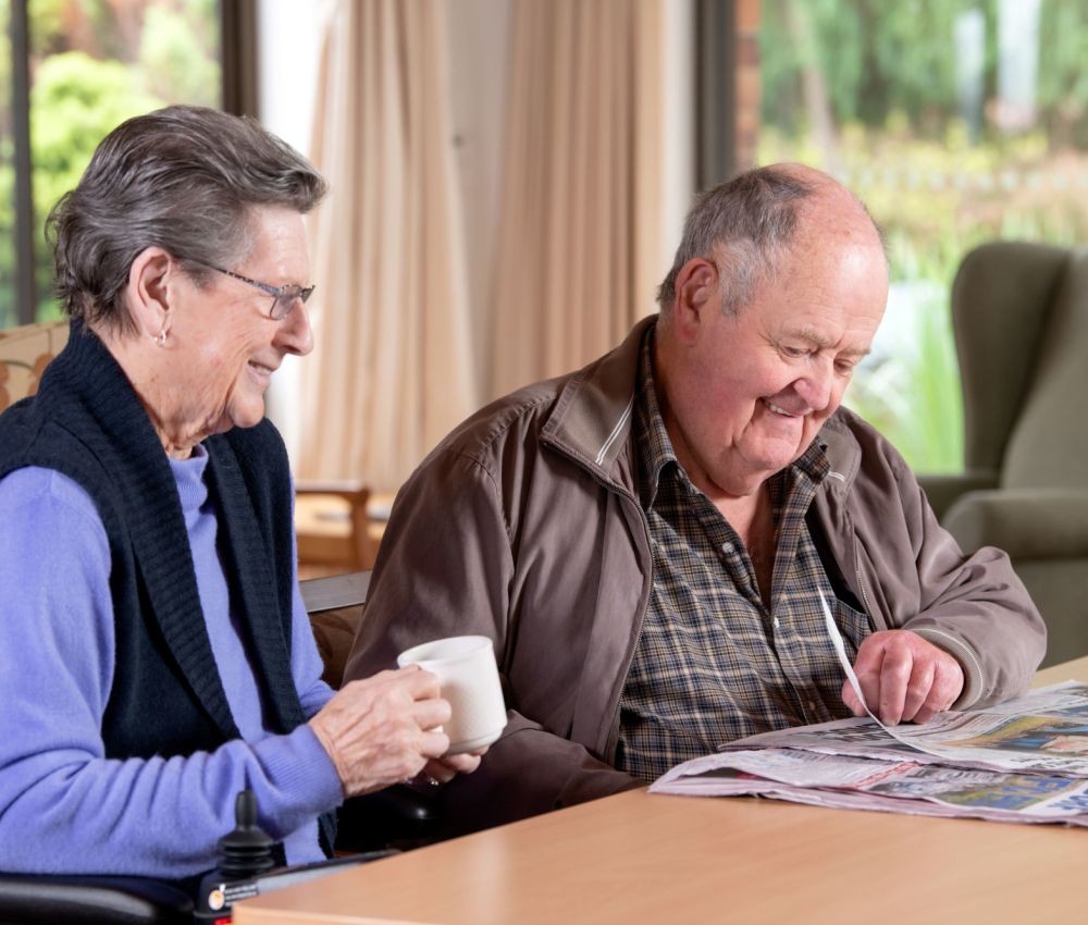 Eldercare Sash Ferguson volunteer smiling at a resident while reading the newspaper to her.