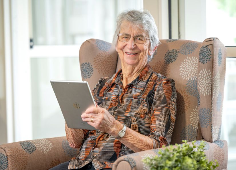 Eldercare resident sitting in an armchair, holding an iPad and smiling at the camera.