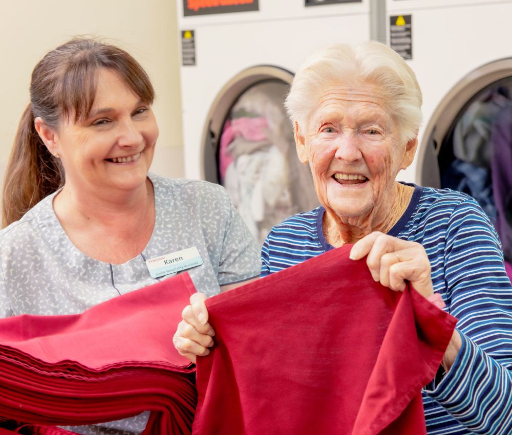 Eldercare Seaford staff member Karen with resident Alice smiling at the camera while folding red napkins in the laundry.