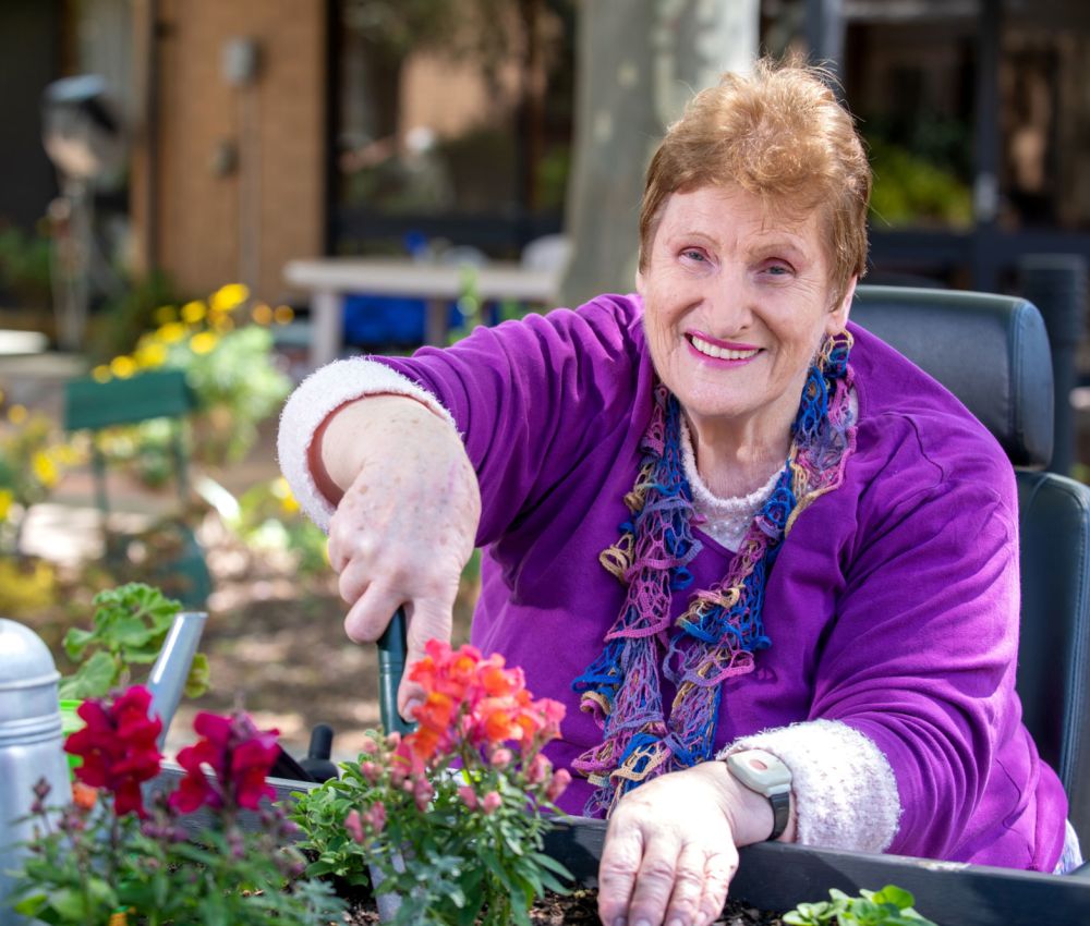 Eldercare Acacia Court resident Bonnie smiling at the camera while tending to some colourful flowers in a raised garden bed.