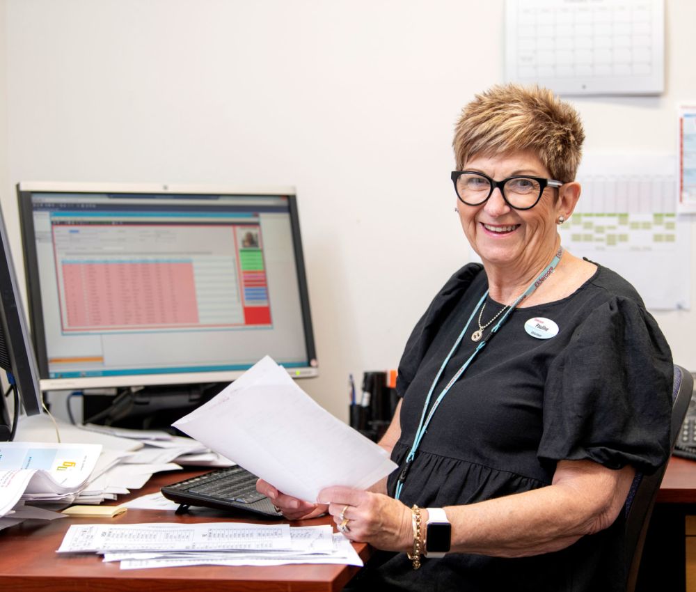 Eldercare The Lodge volunteer Pauline sitting at a desk with some paperwork while smiling at the camera.