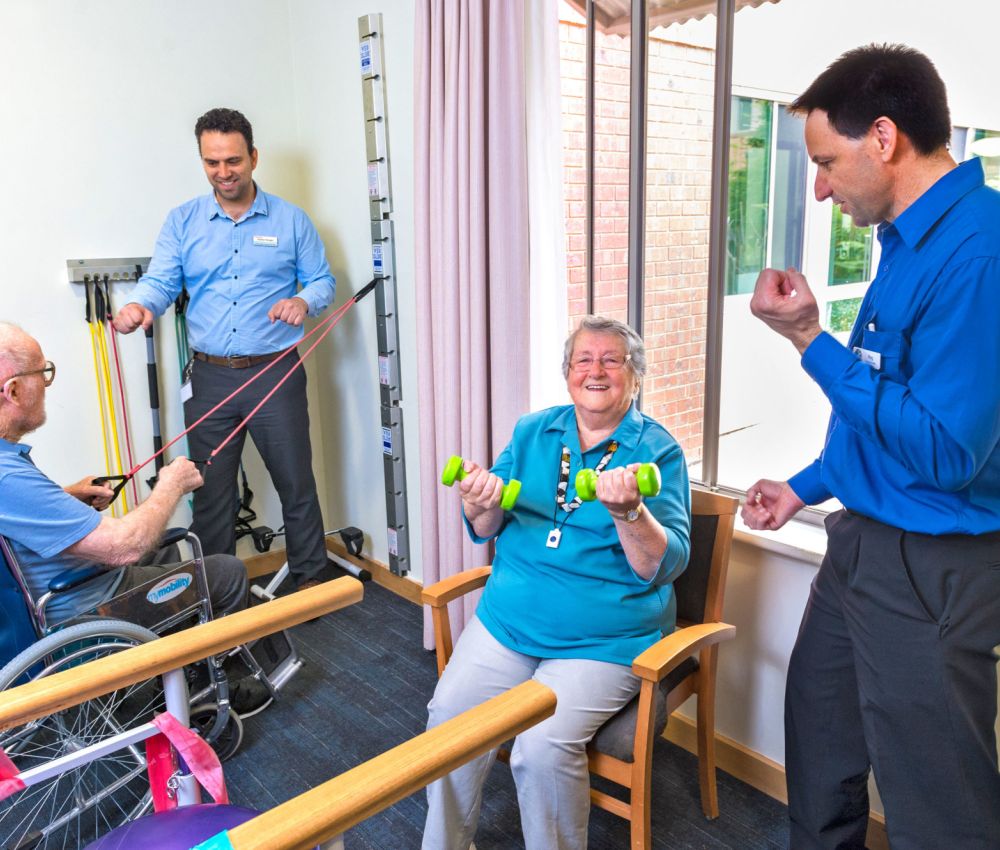 Eldercare allied health staff members instructing two seated residents to use gym equipment.