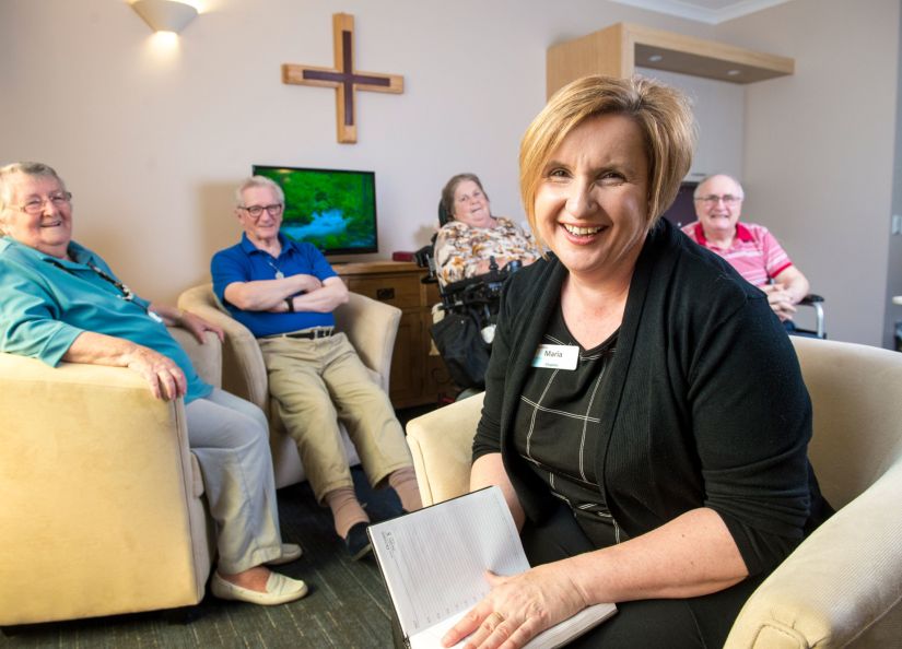 Eldercare Evanston Park Chaplain Maria smiling at the camera with four residents smiling while seated in a group behind her.