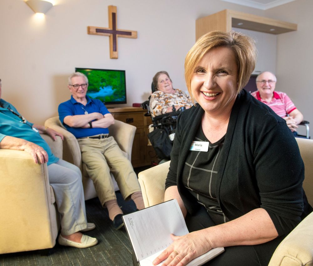 Eldercare Evanston Park Chaplain Maria smiling at the camera with four residents smiling while seated in a group behind her.