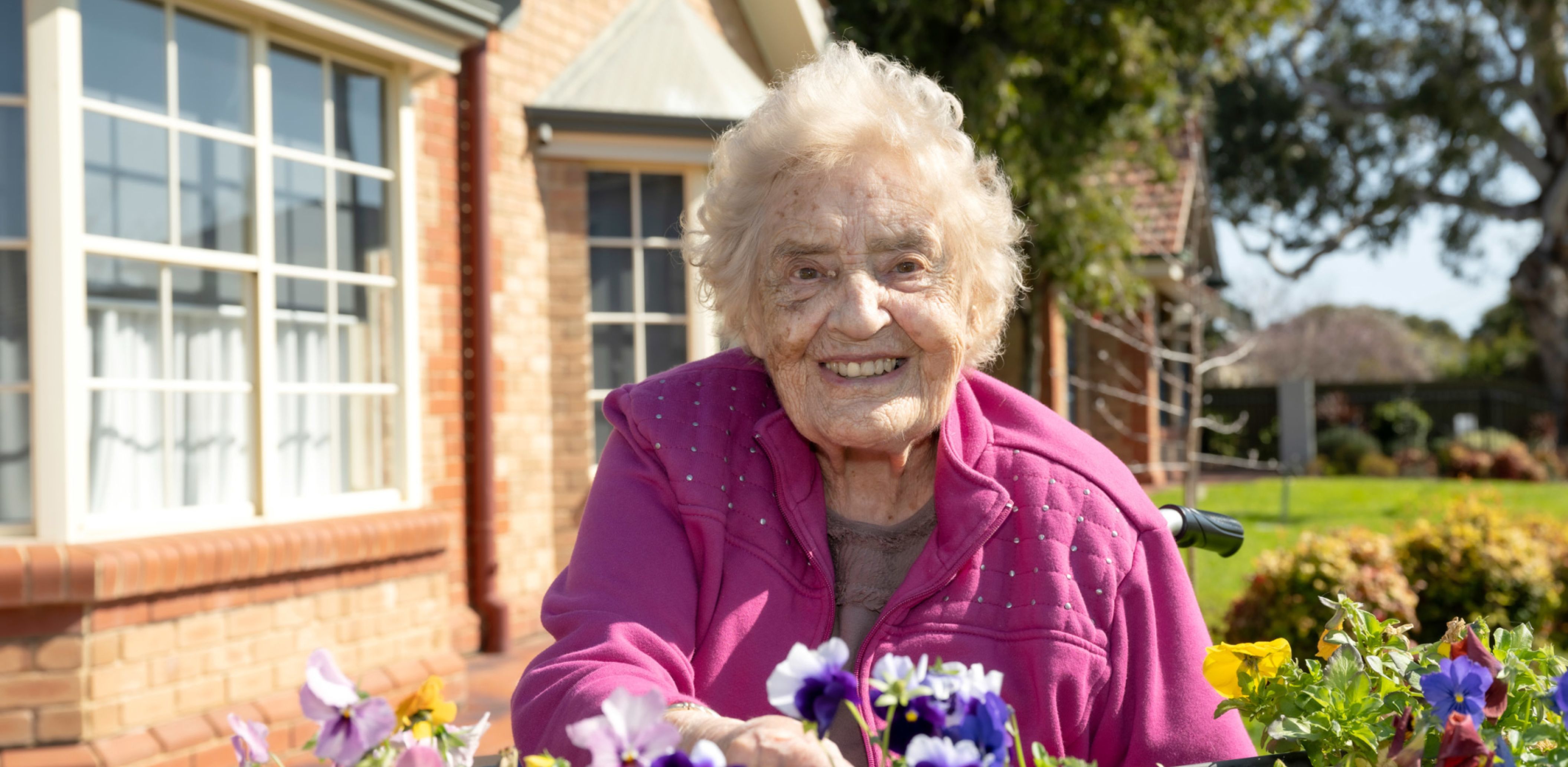 An Eldercare resident smiles at the camera while sitting by a raised garden bed at an aged care home.