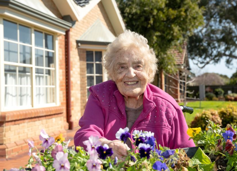 An Eldercare resident smiles at the camera while sitting by a raised garden bed at an aged care home.