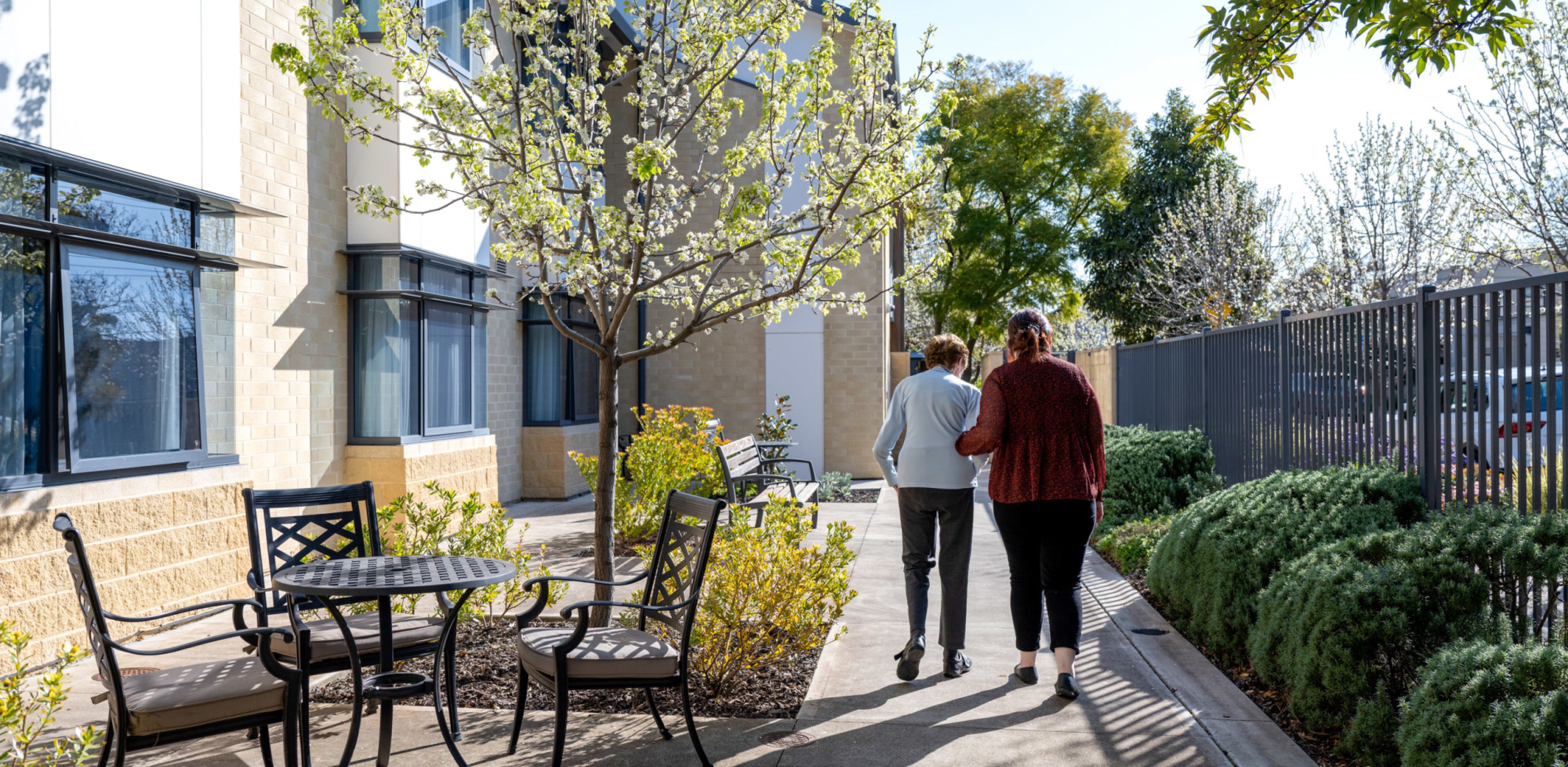 An Eldercare resident and volunteer walk through a sun-soaked courtyard at an aged care home.