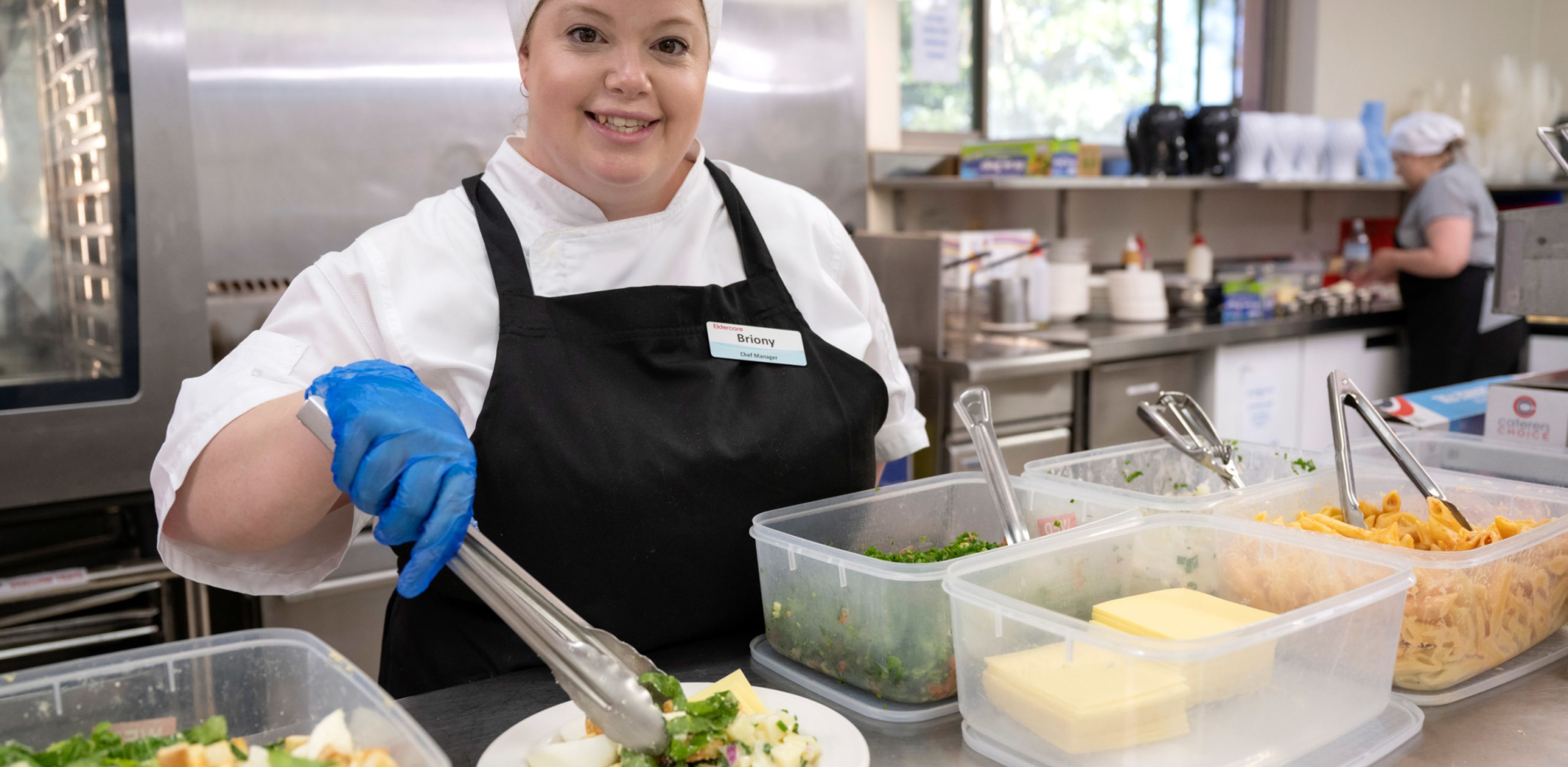 An Eldercare chef manager smiles at the camera and prepares a salad plate in the kitchen at an aged care home.
