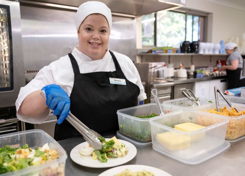 An Eldercare chef manager smiles at the camera and prepares a salad plate in the kitchen at an aged care home.