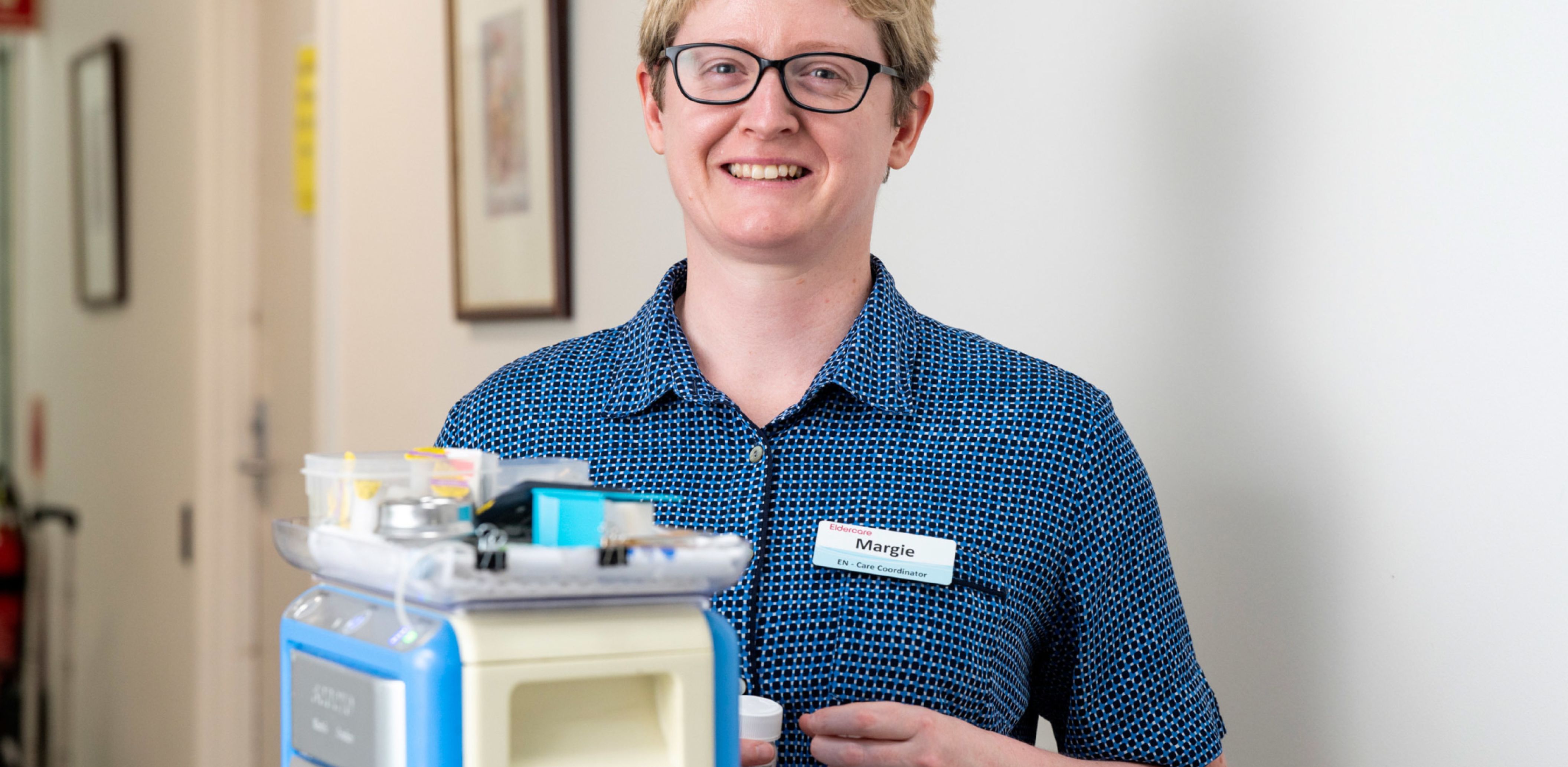 Eldercare Enrolled Nurse smiling at the camera from behind a medication trolley.