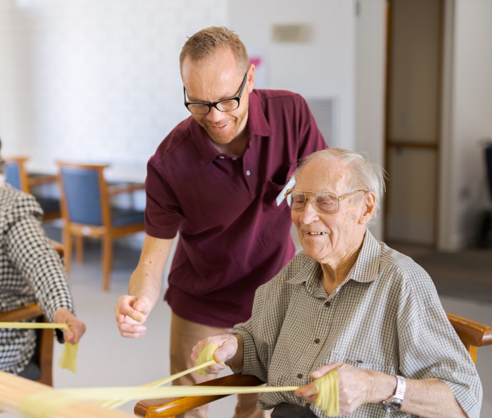 An Eldercare physiotherapist stands by a resident who is sitting at a set of parallel bars and using a resistance band to stretch his arms.