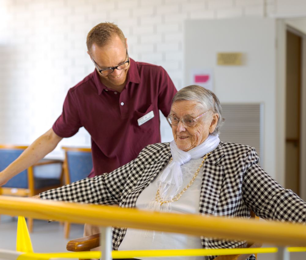 An Eldercare physiotherapist stands by a resident who is sitting at a set of parallel bars and using a resistance band to stretch her arms.