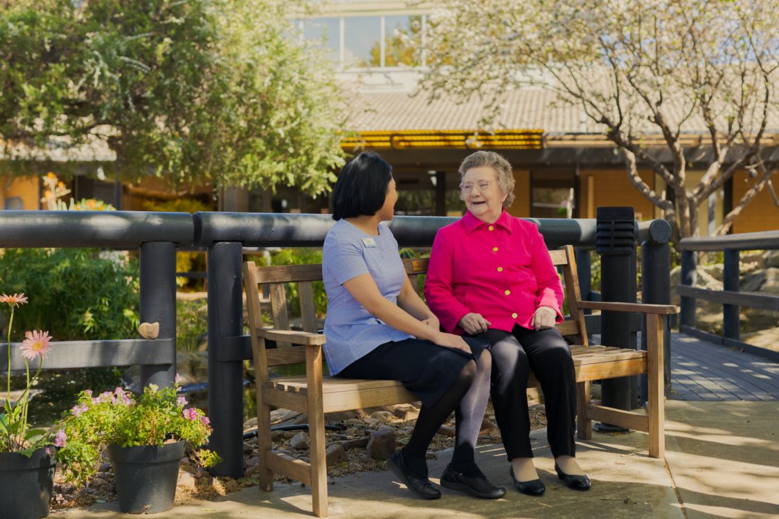An Eldercare staff member sits with a resident on a park bench by the duck pond at Acacia Court.