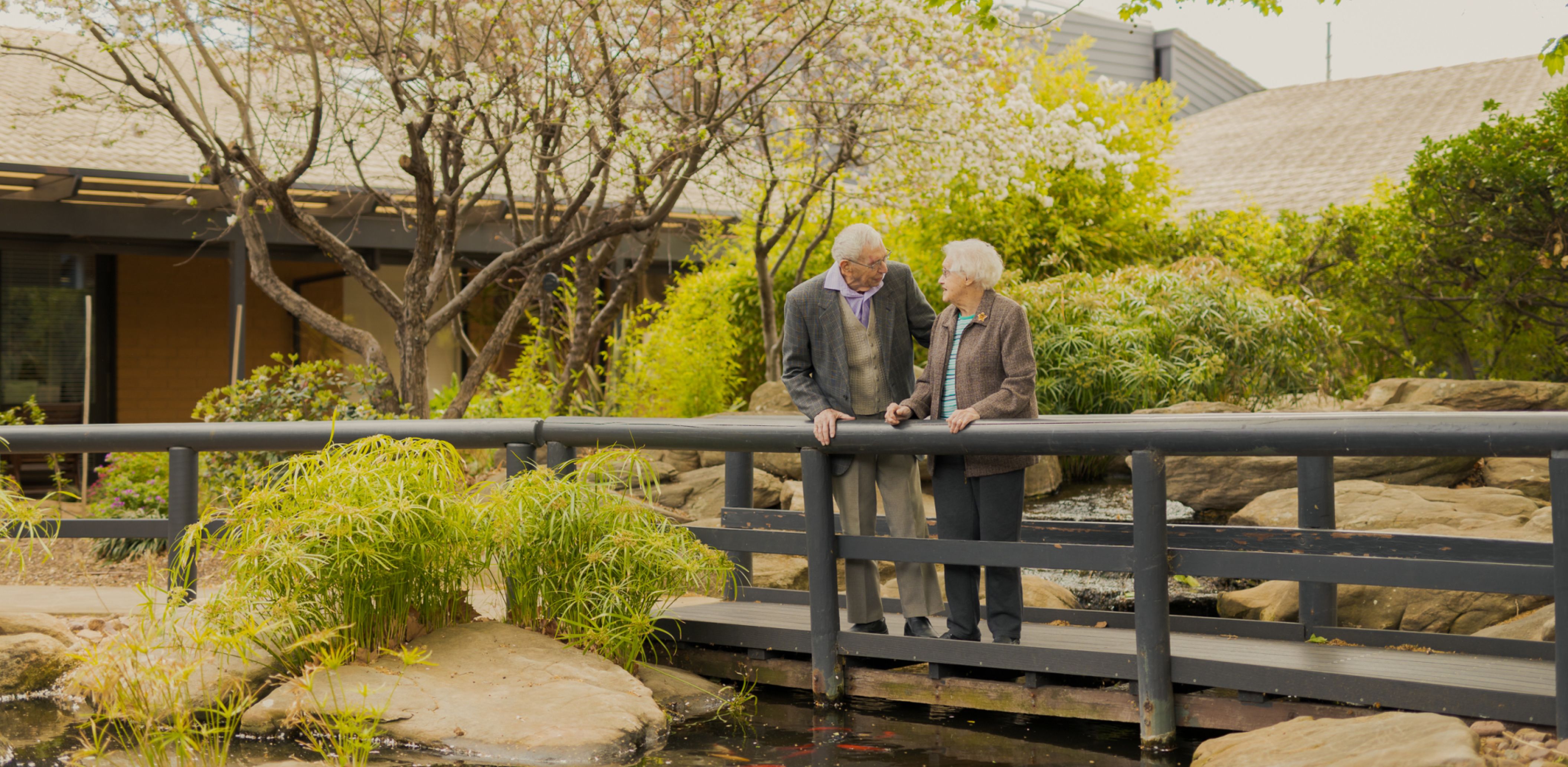 Two Eldercare residents, a couple, smile at each other while on a bridge over the duck pond at Acacia Court.