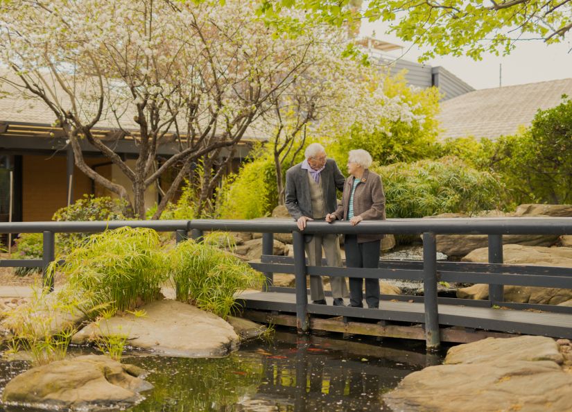 Two Eldercare residents, a couple, smile at each other while on a bridge over the duck pond at Acacia Court.