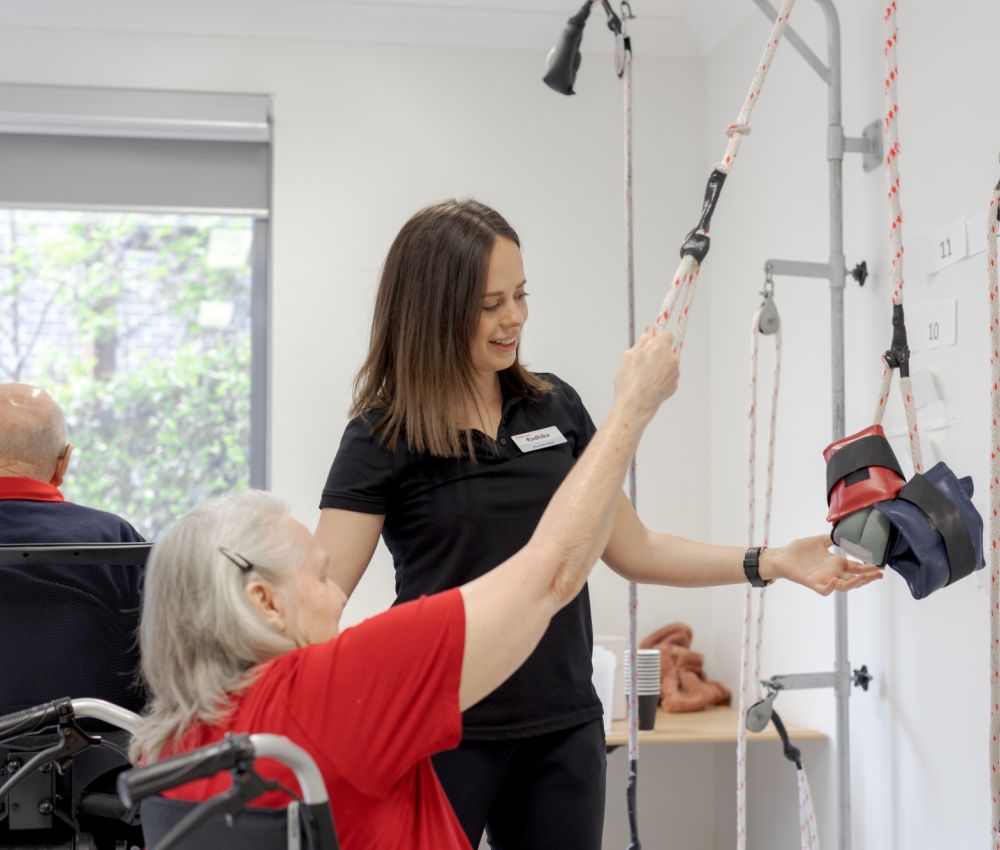 An Eldercare physiotherapist stands by a resident as she sits and uses a pulley machine in the gym.