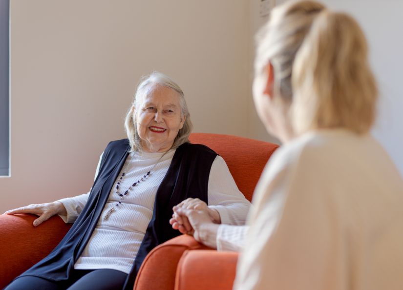 An Eldercare resident smiles at her daughter and holds her hand while sitting in an armchair.