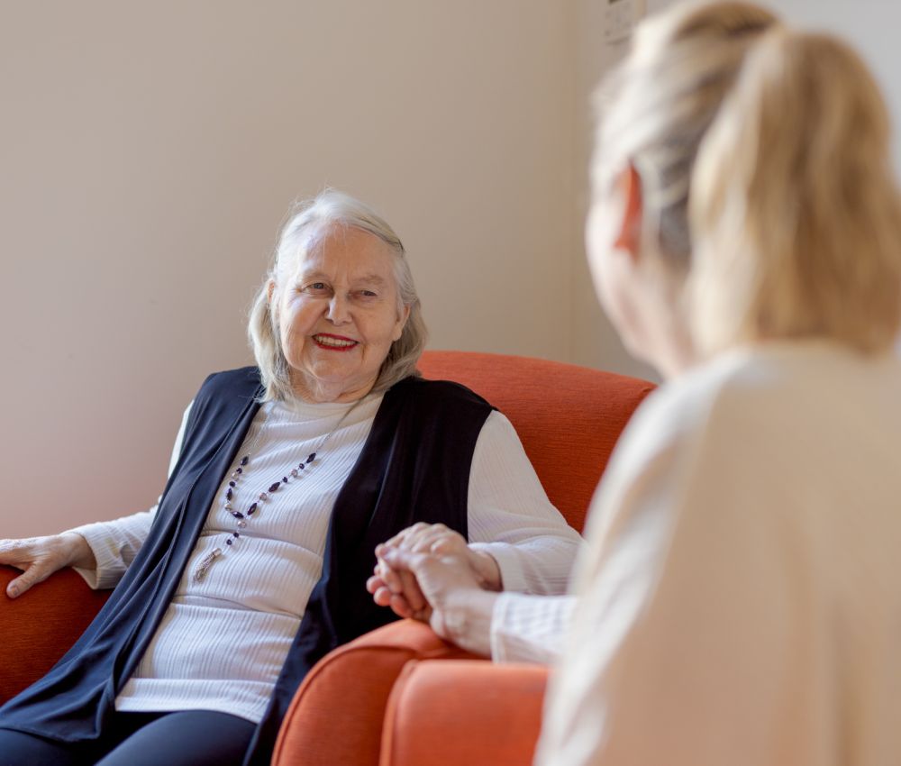 An Eldercare resident smiles at her daughter and holds her hand while sitting in an armchair.