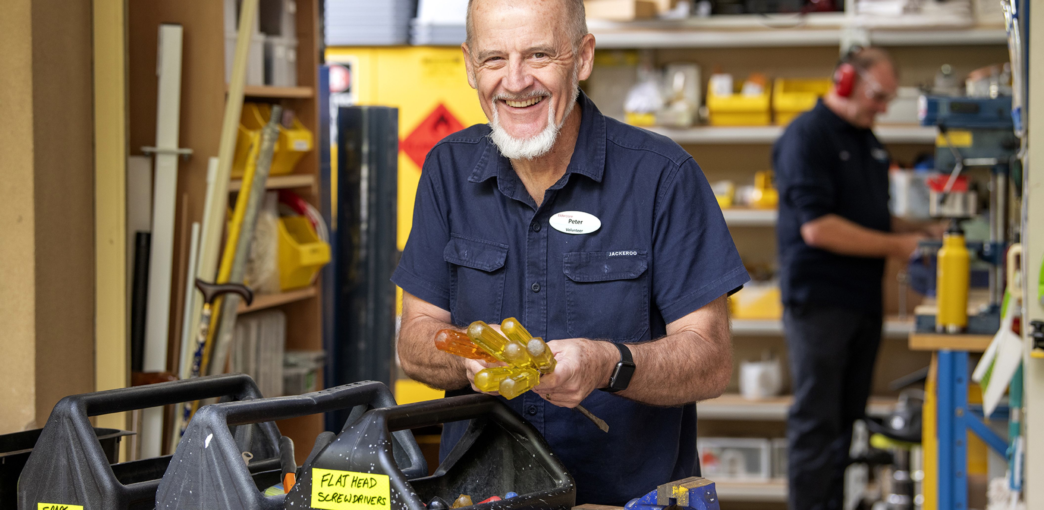 An Eldercare volunteer smiles at the camera and sorts some tools while a Property Services Officer uses a circular saw in the workshop.