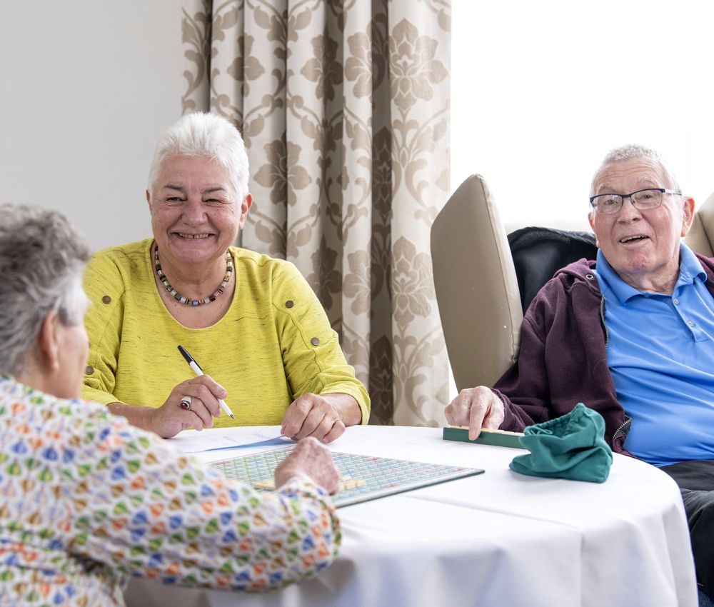 An Eldercare volunteer plays Scrabble with two Eldercare residents while smiling.