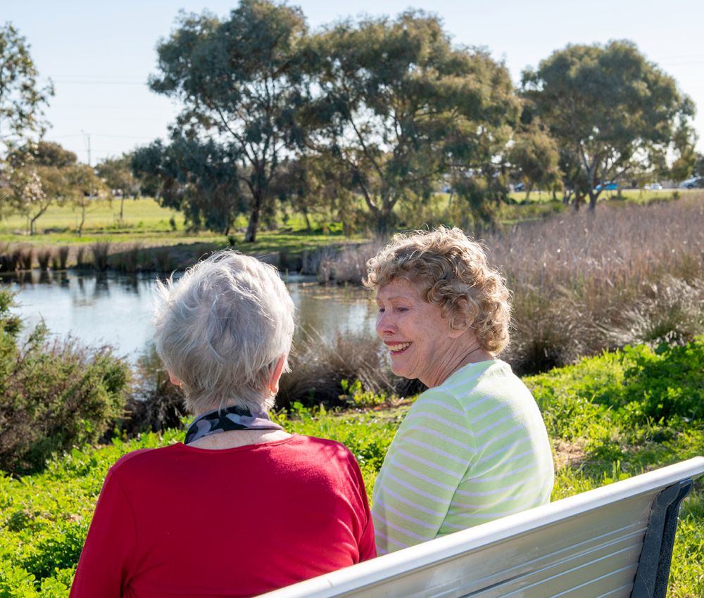 Two residents sit together outdoors on a park bench by a lake near Eldercare Seaford.
